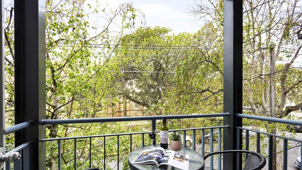 A balcony with a glass table, two wicker chairs, a wine bottle, and a magazine. - Quest St Kilda Bayside (St Kilda)