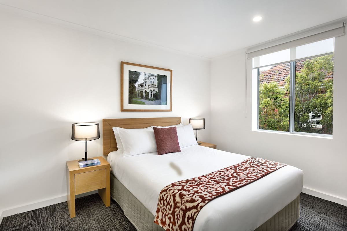 A hotel room with a large bed, two bedside tables with lamps, a framed picture on the wall, and a window with a view of trees.