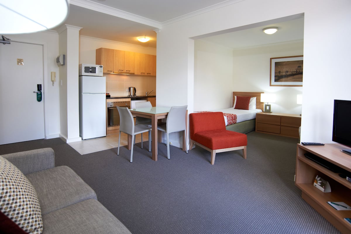 A compact hotel room with a kitchenette, a dining area, a sofa, and a TV.