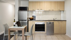 A modern kitchen with wooden cabinets, a built-in microwave, a dishwasher, and a dining area with a laptop and wine bottle. - Quest St Kilda Bayside (St Kilda)