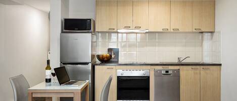 A modern kitchen with wooden cabinets, a built-in microwave, a dishwasher, and a dining area with a laptop and wine bottle.