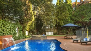 A swimming pool with a waterfall feature, surrounded by lush greenery and a patio area with lounge chairs and a table. - Quest St Kilda Bayside (St Kilda)