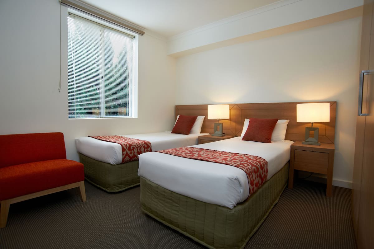 A hotel room with two beds, a red sofa, and a window with blinds.
