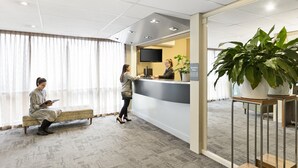 A reception area with a reception desk, a bench, a television, and a large potted plant. - Quest St Kilda Bayside (St Kilda)