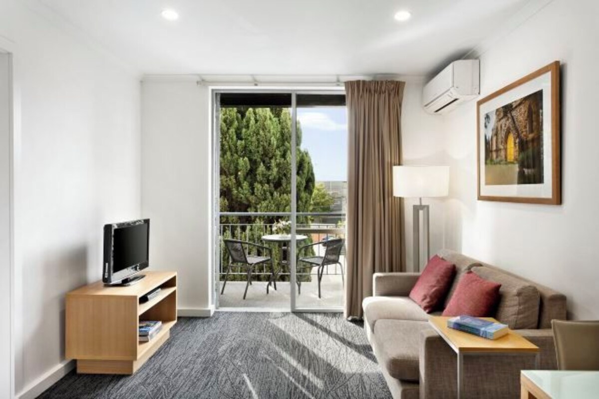 A hotel room with a TV on a wooden stand, a sofa with red cushions, a table with a lamp, and a balcony with a view of trees.