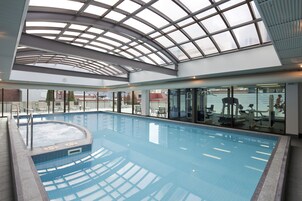 Una piscina interior con techo de vidrio, agua cristalina y equipo de ejercicio al fondo.