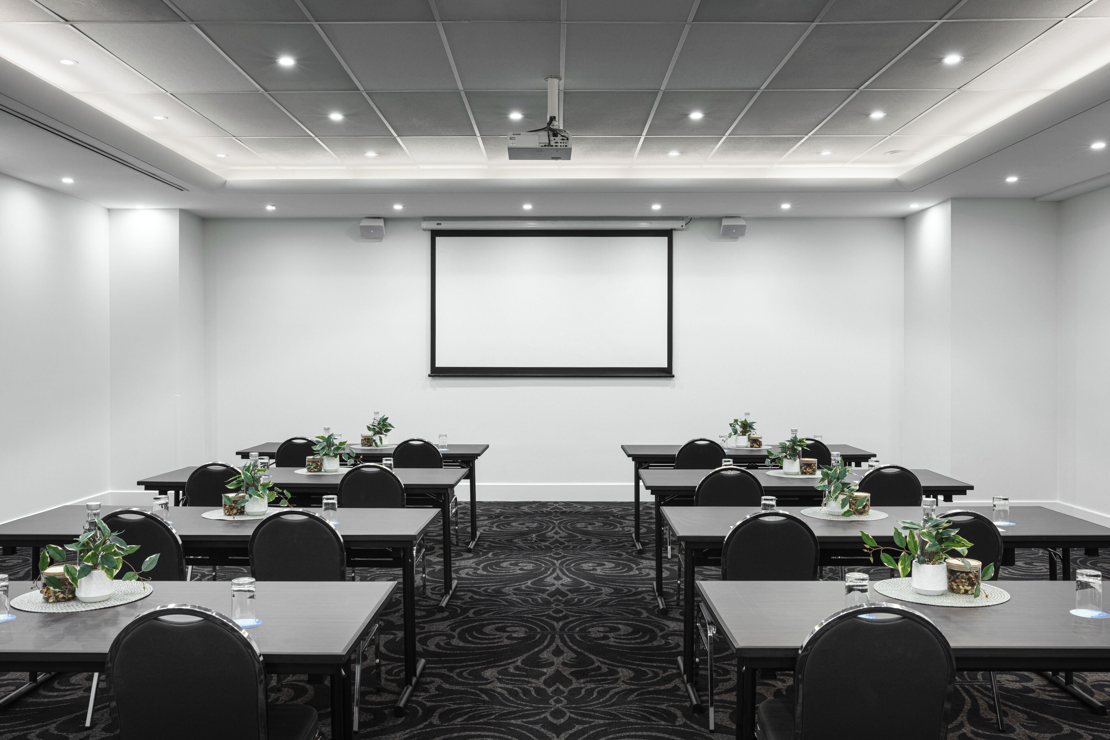 Una sala de conferencias con pantalla de proyector, mesas con sillas y plantas en macetas.