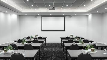 Una sala de conferencias con pantalla de proyector, mesas con sillas y plantas en macetas.