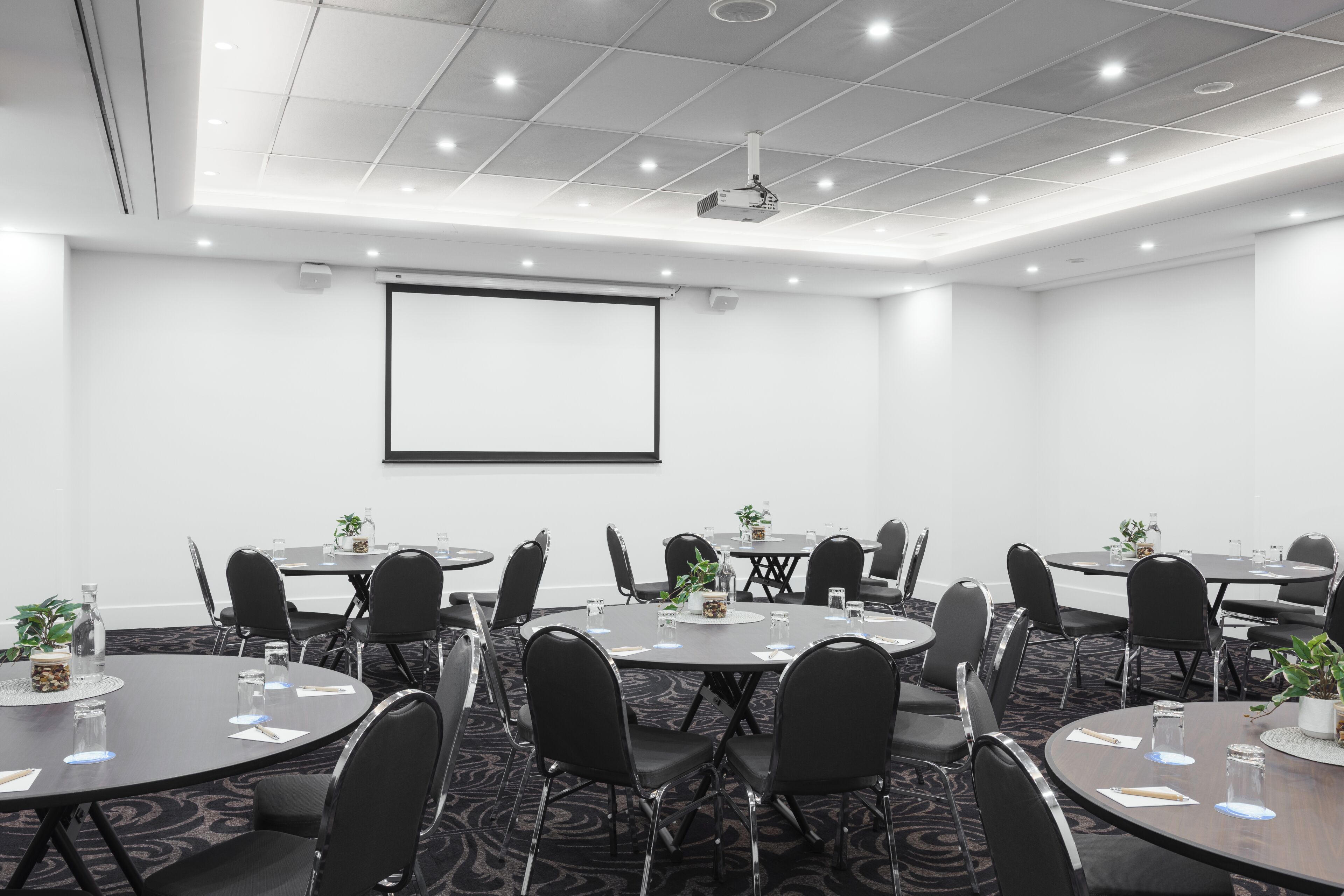 Un salón de conferencias con una pantalla de proyector, varias mesas con sillas y piso alfombrado.
