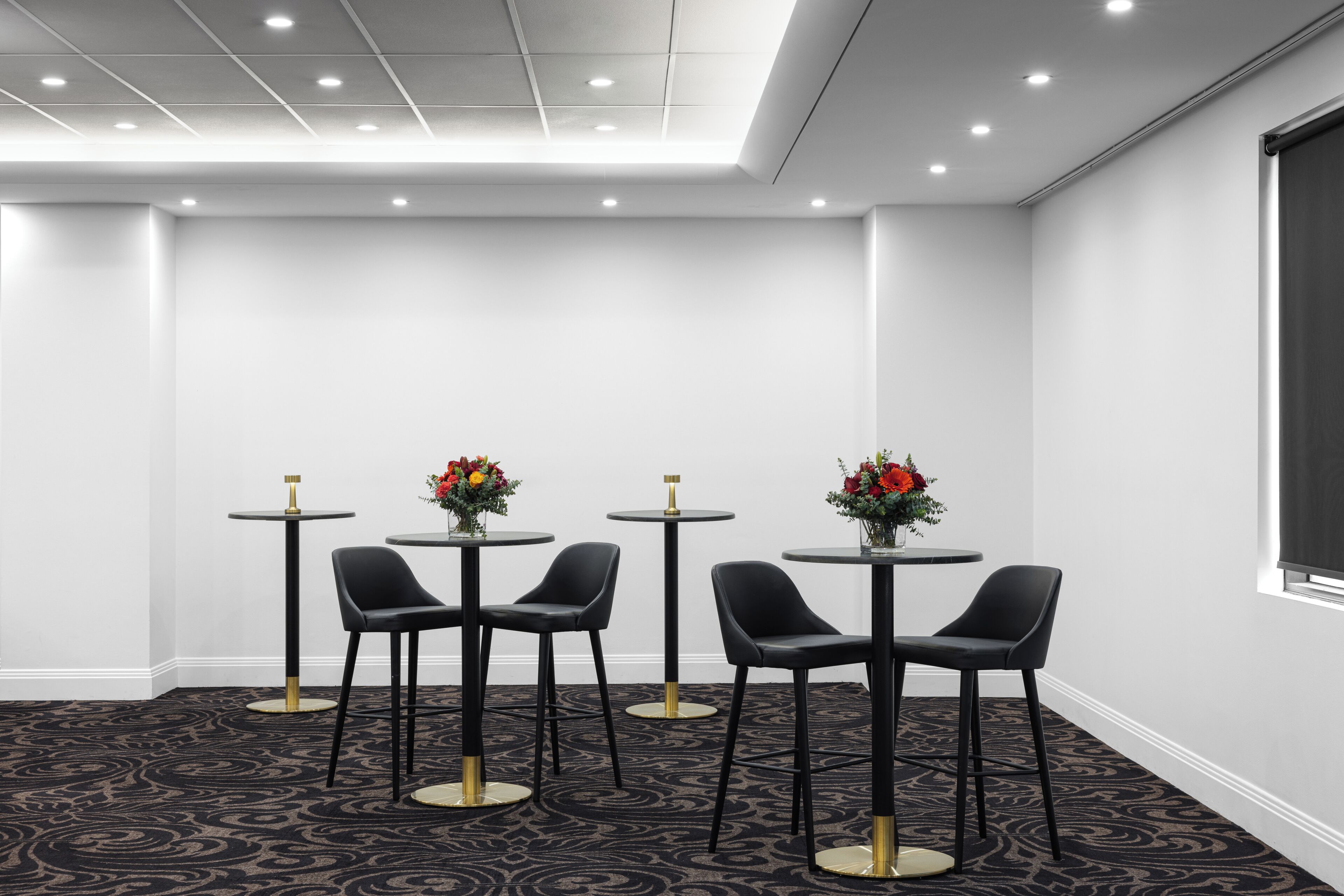 A conference room with four tables, each with a chair and a vase of flowers. The room has a patterned carpet and recessed ceiling lighting.