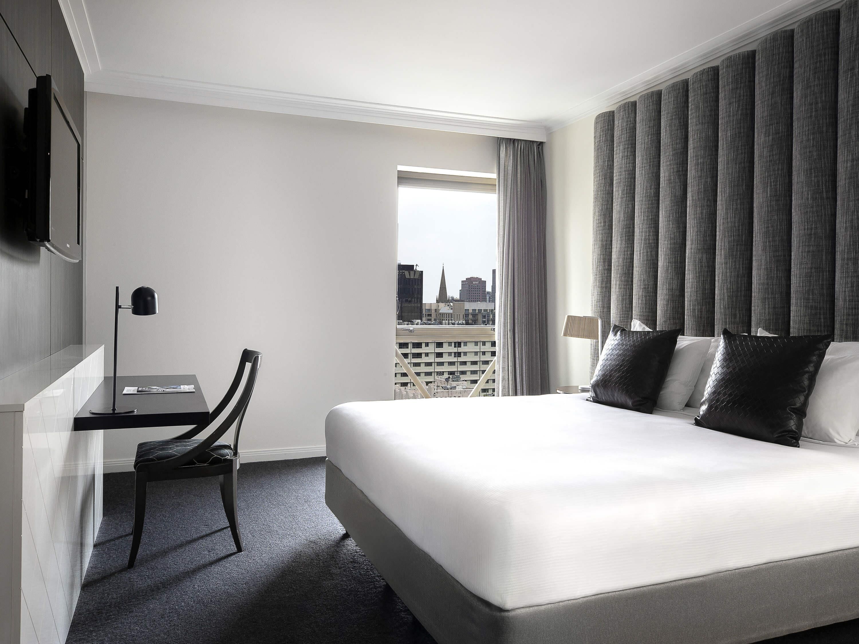 A hotel room with a bed, a desk, a chair, a TV, and a view of the cityscape.