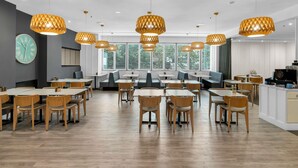 Un espace de restauration moderne avec des tables et des chaises en bois, de grandes fenêtres équipées de stores et des luminaires suspendus.