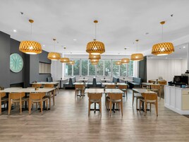 Un comedor moderno con mesas y sillas de madera, ventanas amplias con persianas y lámparas colgantes.