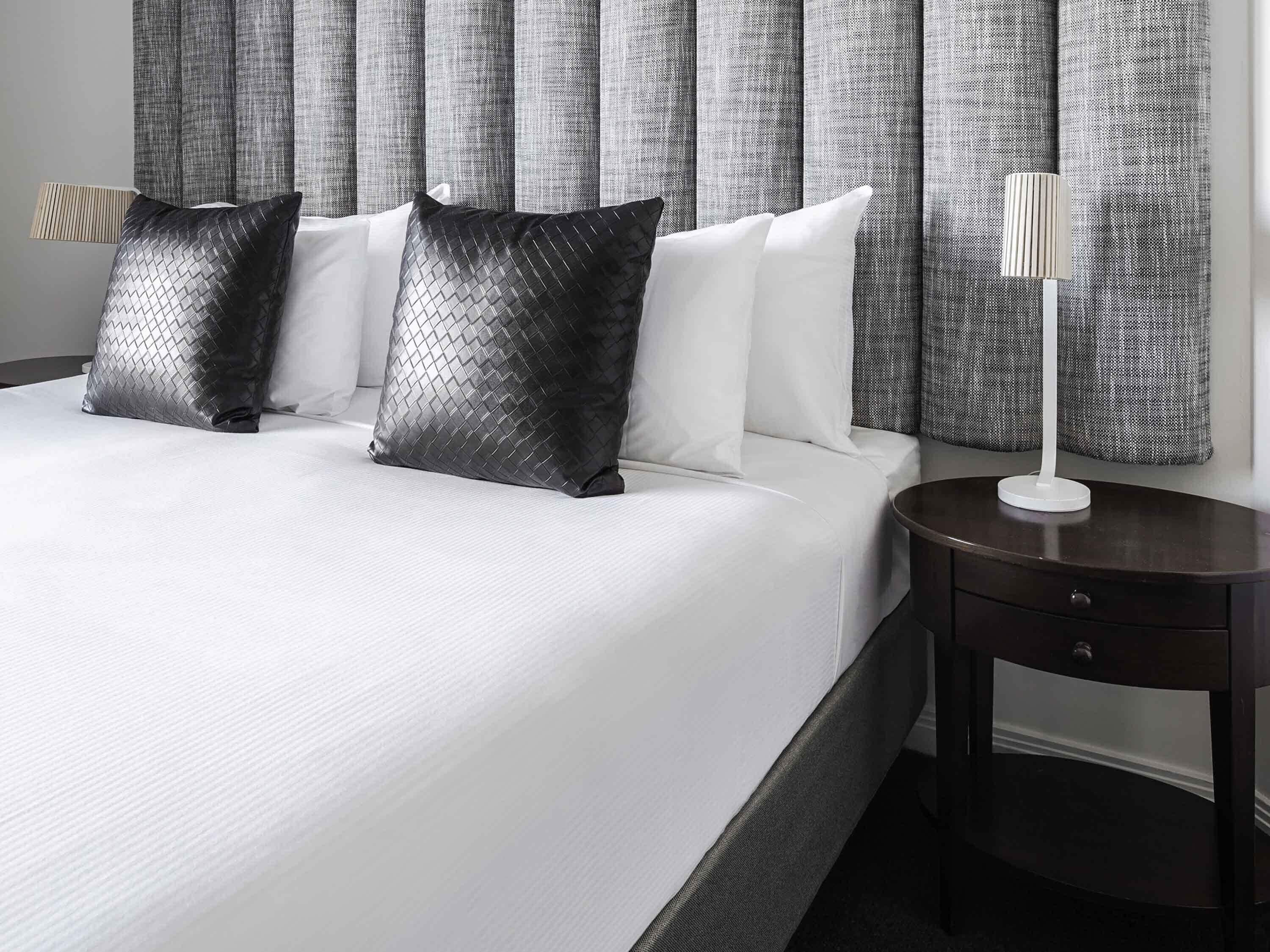 A hotel room with a large bed, two black patterned pillows, white bedding, grey curtains, and a bedside lamp.