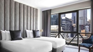 Une chambre d’hôtel avec deux lits, une grande fenêtre donnant sur la ville, une chaise et une lampe.