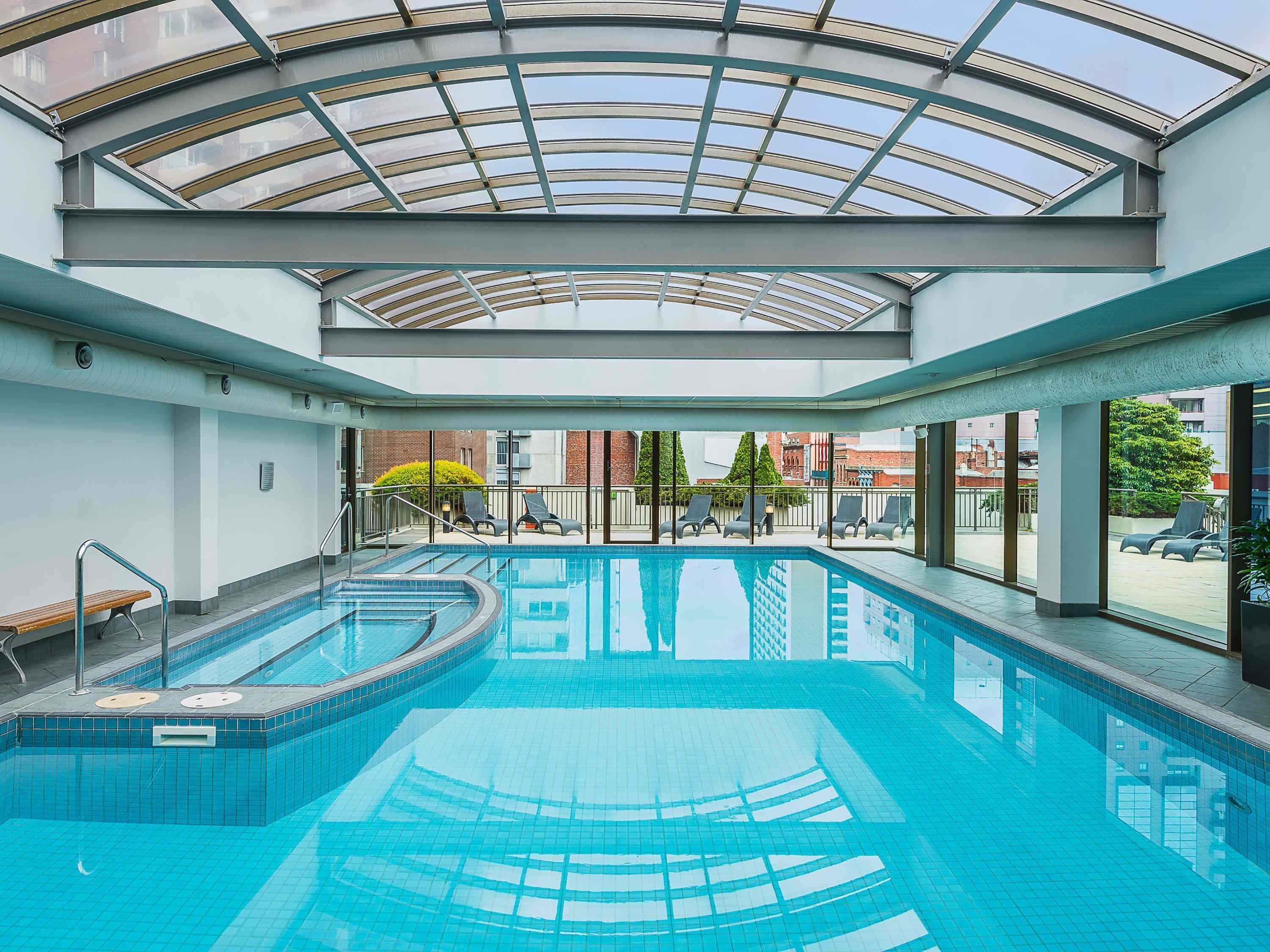 A swimming pool with a glass ceiling, surrounded by lounge chairs and a view of buildings.