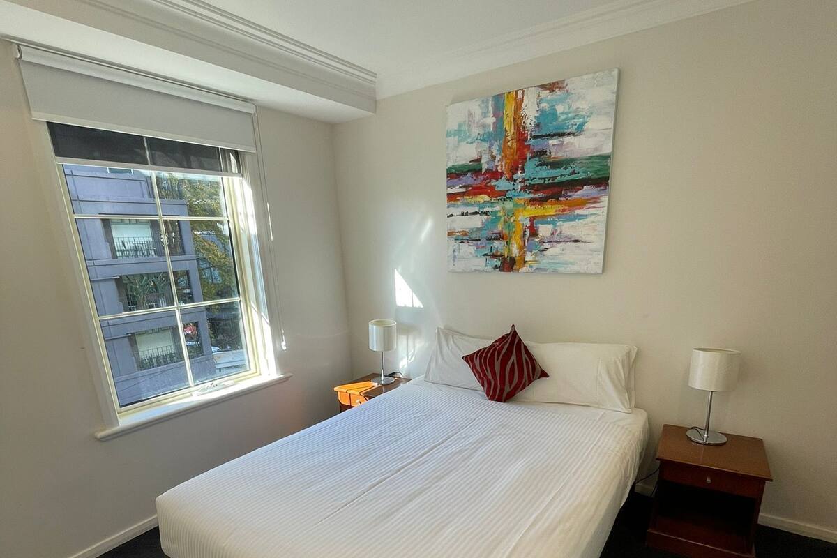 A hotel room with a large bed, a window with blinds, a nightstand with a lamp, and a painting on the wall.
