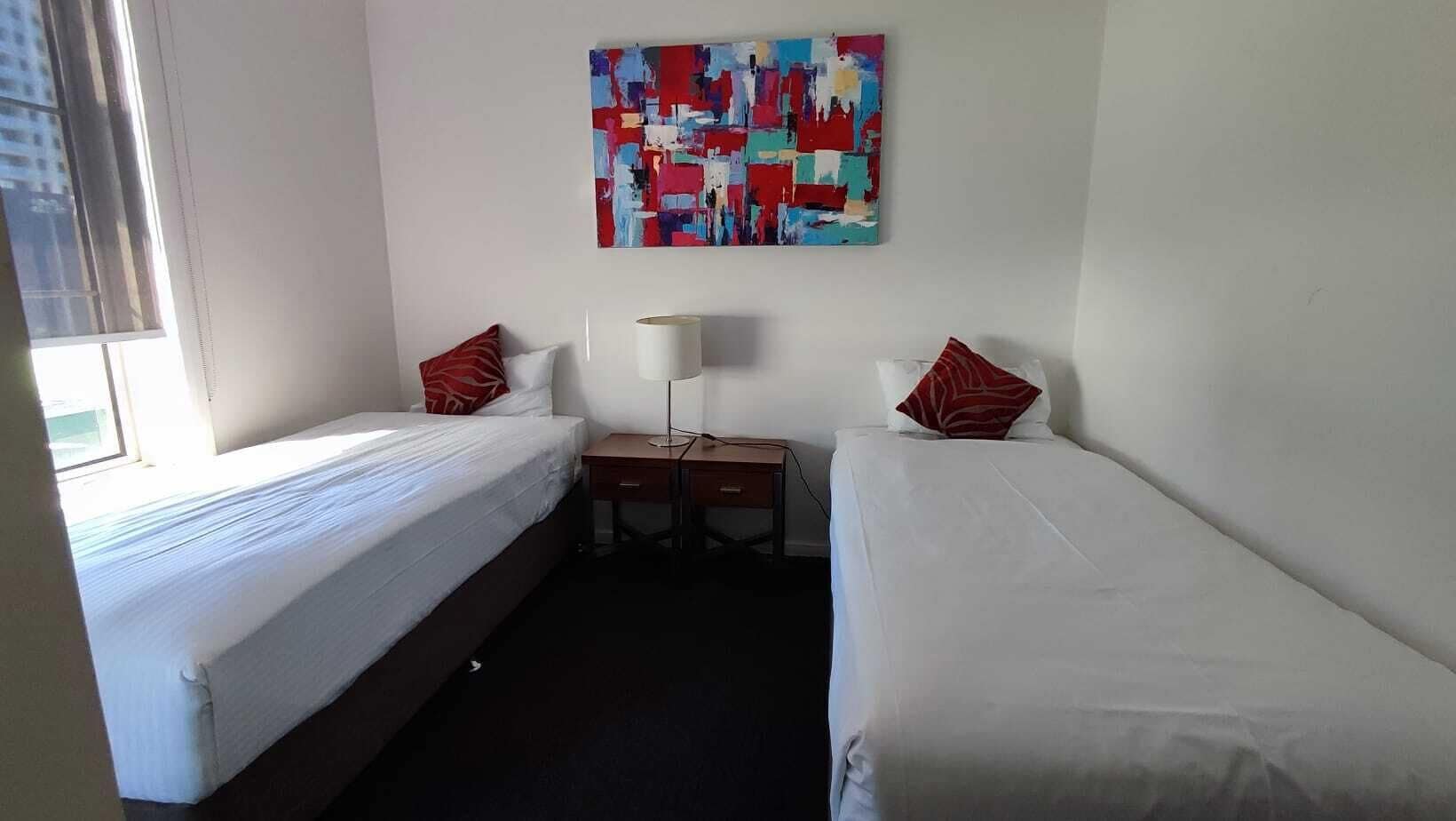 Une chambre d’hôtel avec deux lits, un bureau, une lampe et une peinture abstraite aux couleurs vives accrochée au mur.