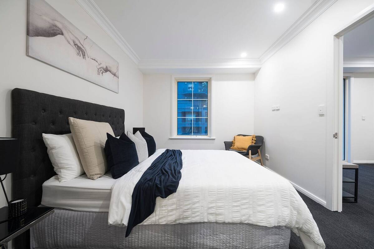 A bedroom with a bed, a nightstand, a chair, a window, and a painting on the wall.