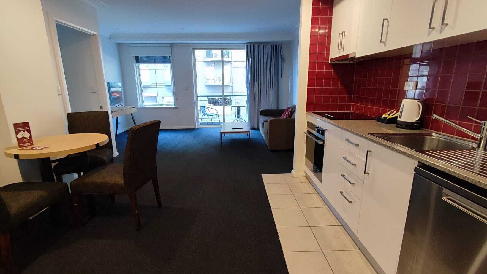 Et moderne køkken med hvide skabe og røde fliser, et spiseområde med et rundt bord og stole, samt udsigt til en balkon med gelænder.