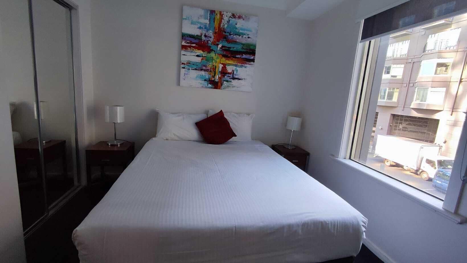 A hotel room with a large bed, two bedside tables, a window with a view of buildings, and a colorful abstract painting on the wall.