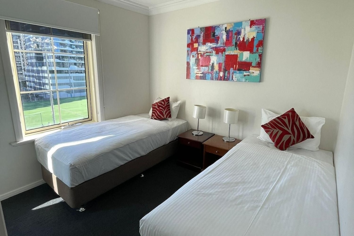 Une chambre d’hôtel avec deux lits, une grande fenêtre donnant sur un court de tennis et une peinture abstraite aux couleurs vives accrochée au mur.