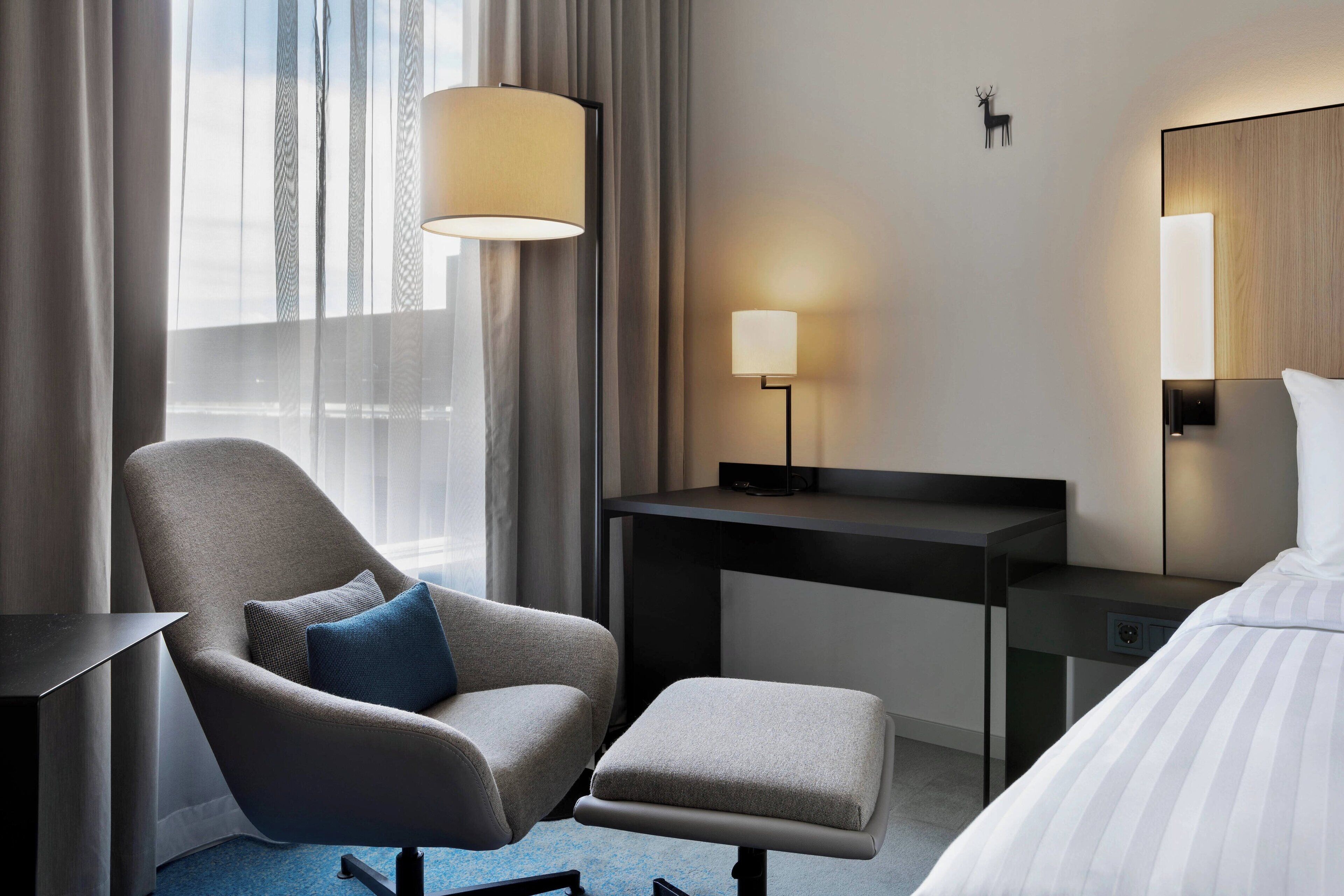 Une chambre d’hôtel moderne avec un fauteuil gris, une petite table, une table de chevet, une lampe, un lit et une grande fenêtre avec des rideaux légers.