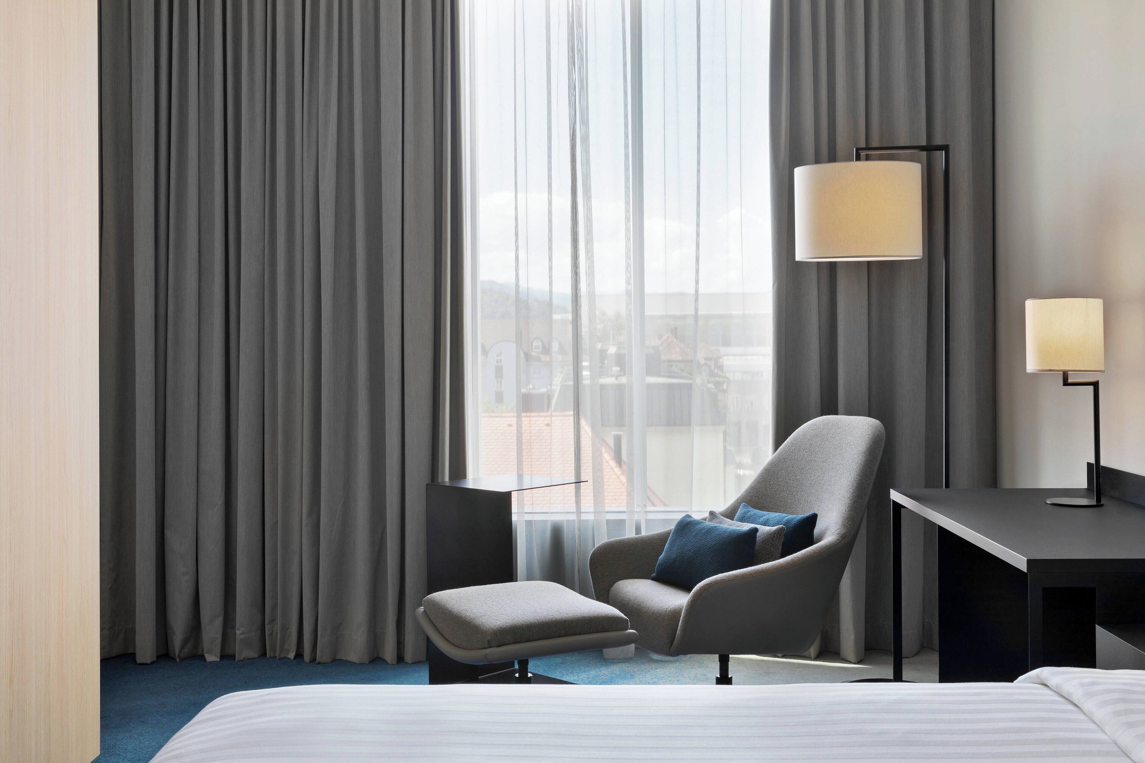 A hotel room with a bed, a chair, a desk, and a lamp.
