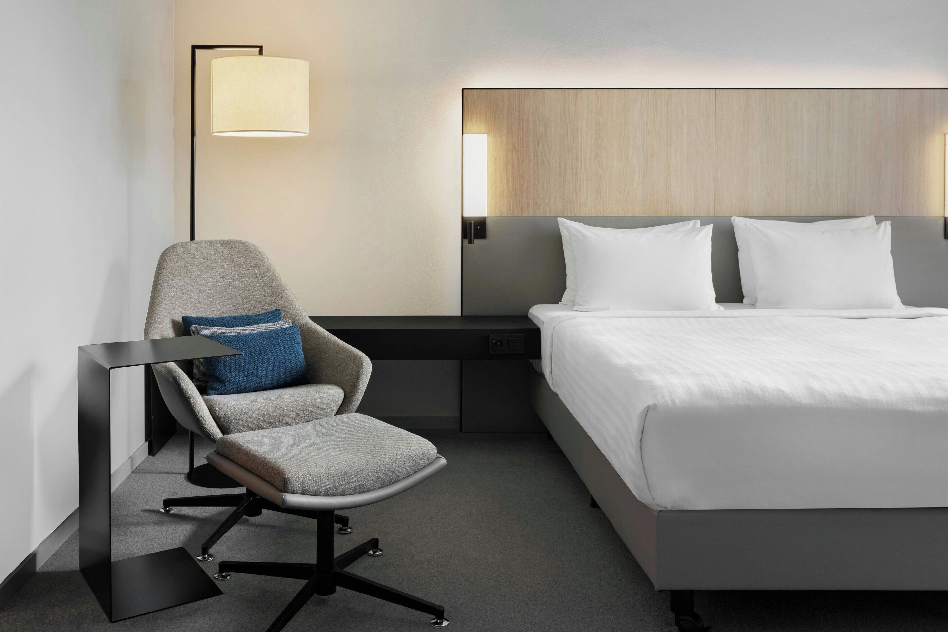 A modern hotel room with a bed, a desk, a chair, and a lamp.