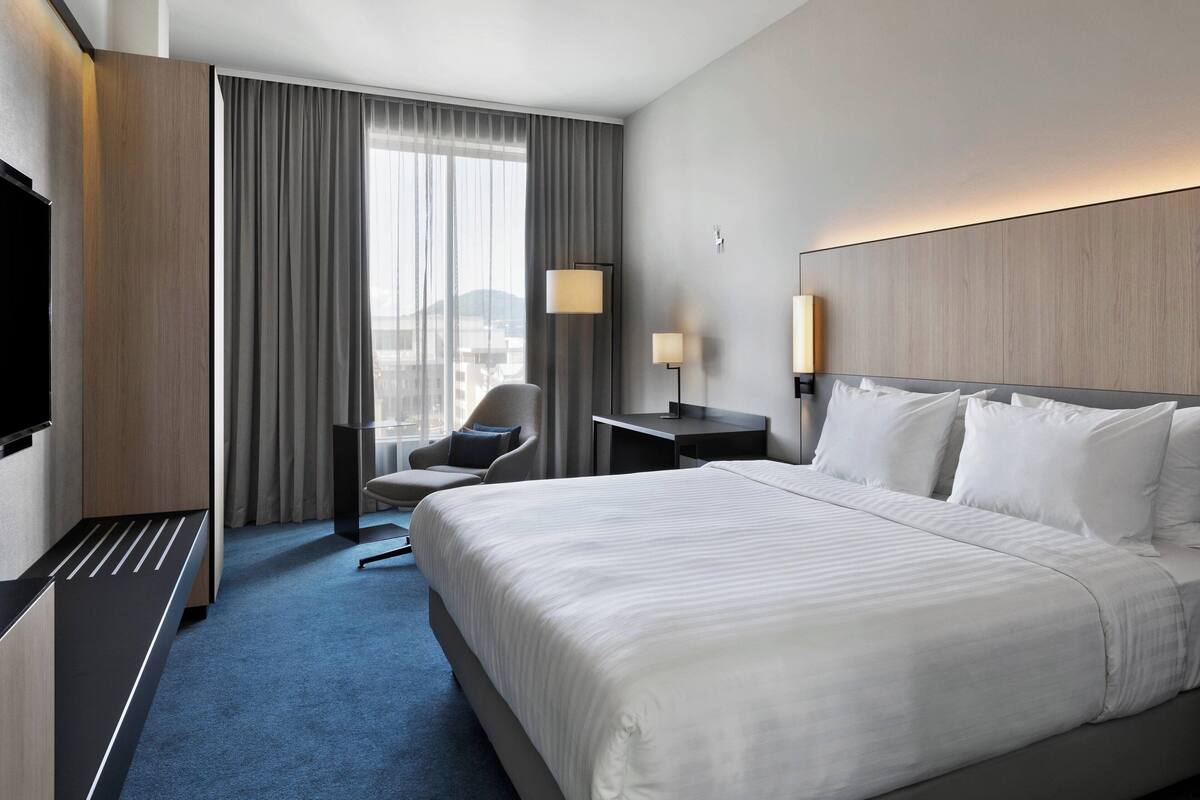 Une chambre d’hôtel avec un grand lit, un bureau, une chaise, une télévision et une fenêtre avec des rideaux.