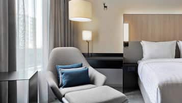 A hotel room with a grey armchair, a bed, a nightstand, a lamp, and a window with curtains.