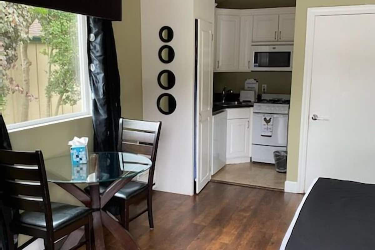 A compact room with a dining table and chairs, a bed, a kitchen area with cabinets and appliances, and a window with curtains.