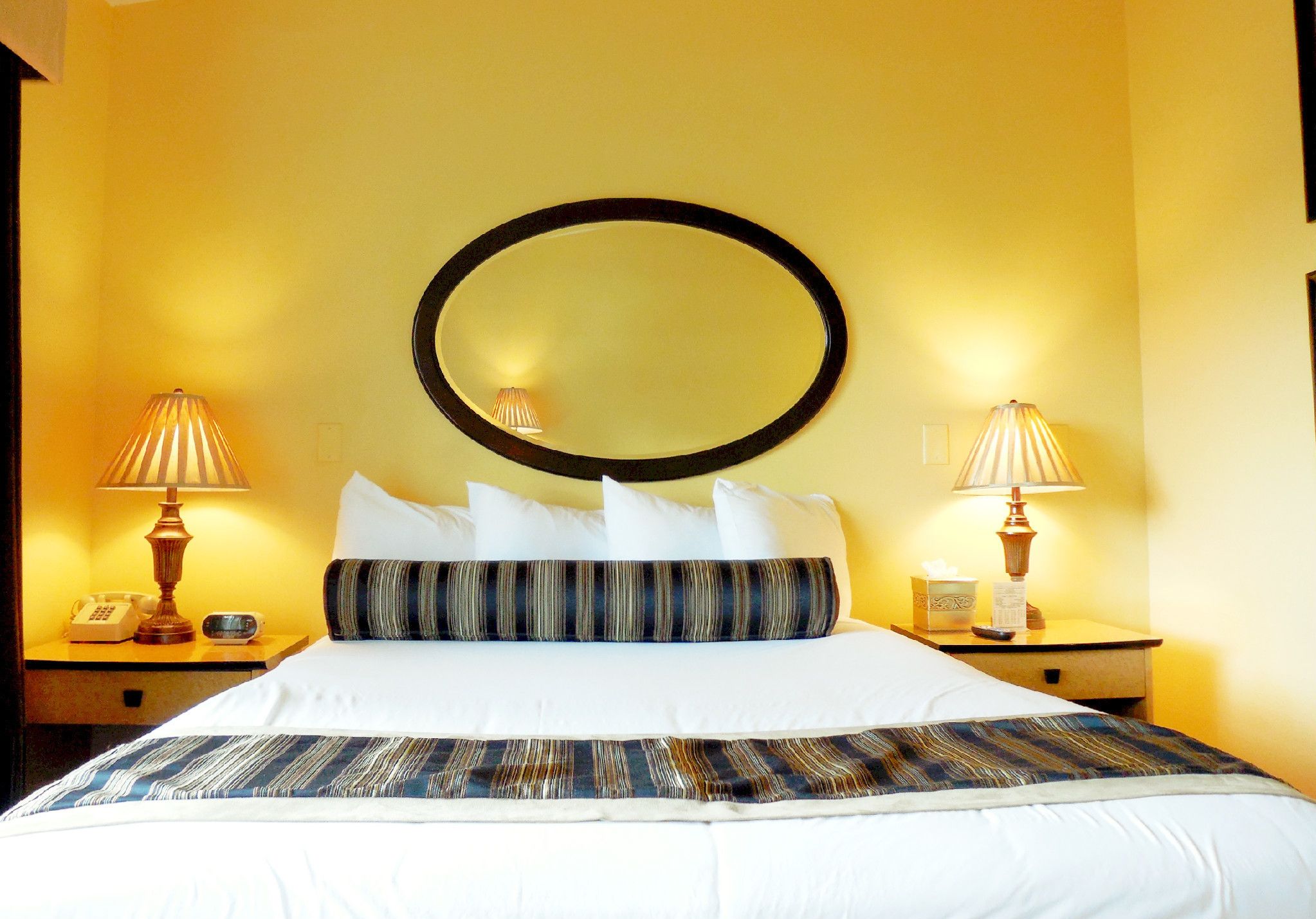 Una habitación de hotel con una cama grande, dos lámparas de noche, un espejo redondo sobre la cama y un teléfono en la mesita.