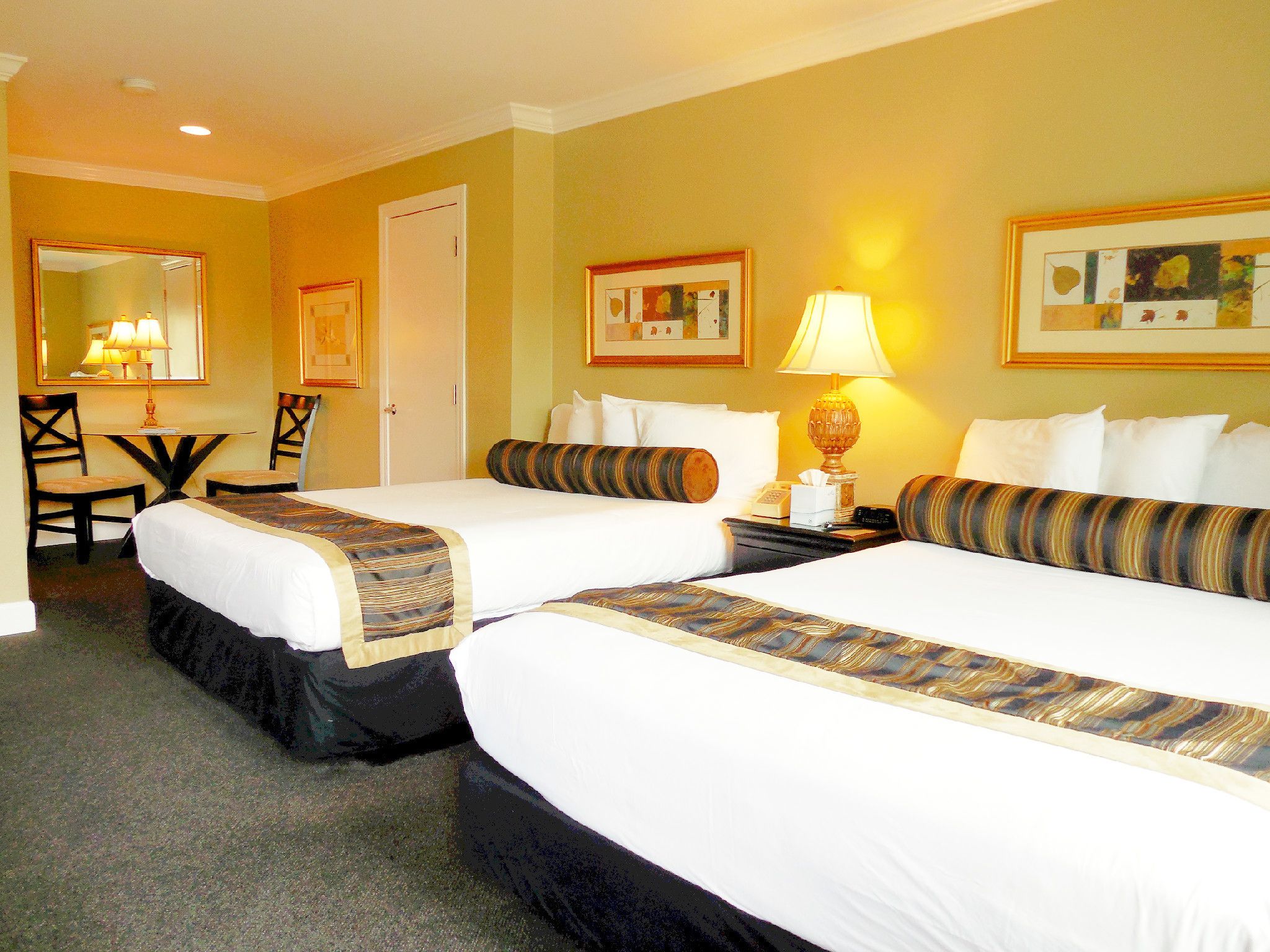 Quarto de hotel com duas camas, uma escrivaninha, cadeiras, abajur e quadros nas paredes.
