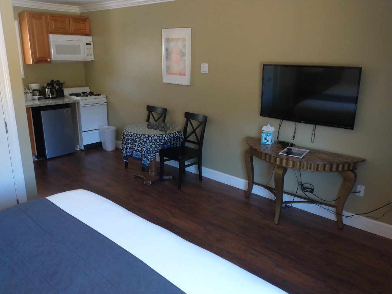 A room with a bed, a dining table with chairs, a kitchen area with a microwave, stove, and refrigerator, and a TV mounted on the wall.