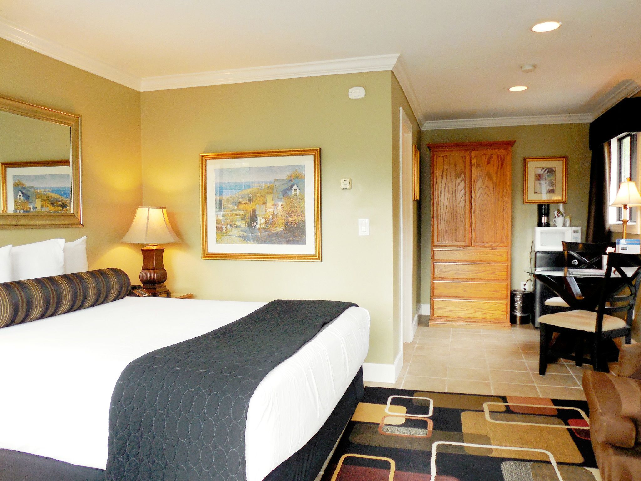 A hotel room with a large bed, a desk, and a chair. There is a painting on the wall and a window with curtains.