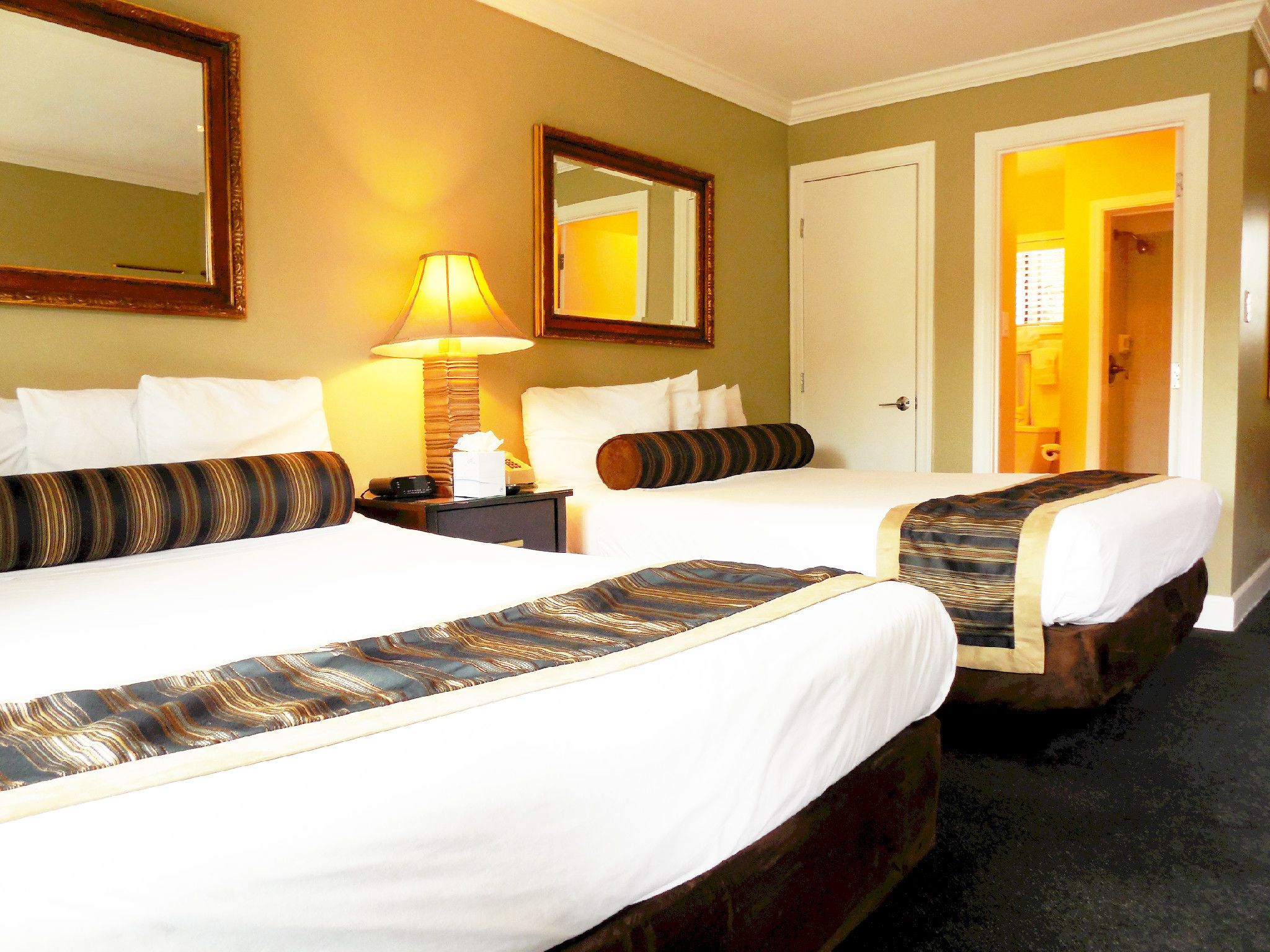 Habitación de hotel con dos camas, una mesita de noche con lámpara, un espejo y una puerta que da a otra habitación.