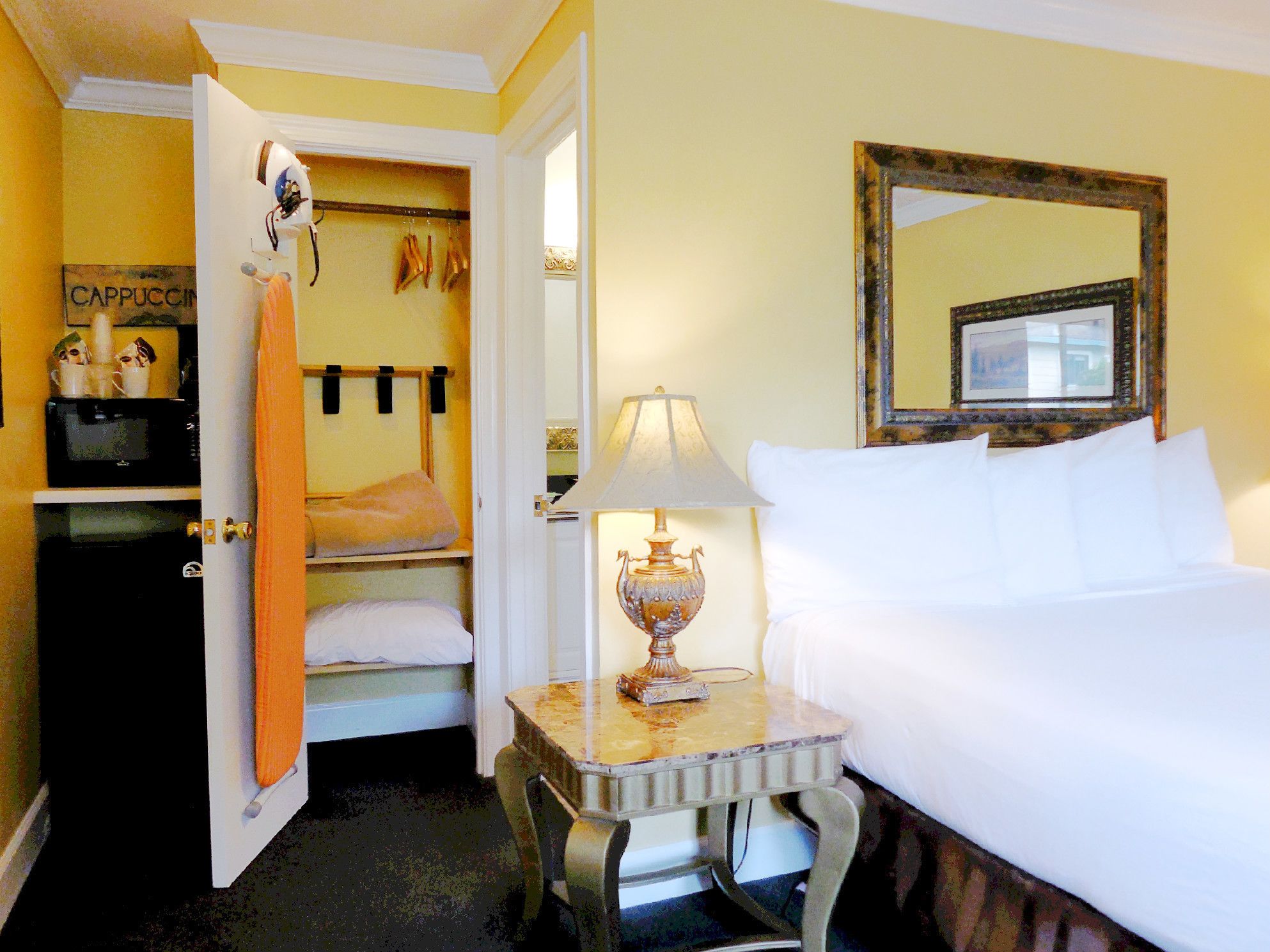A hotel room with a bed, a table with a lamp, a wardrobe, and a mirror on the wall.