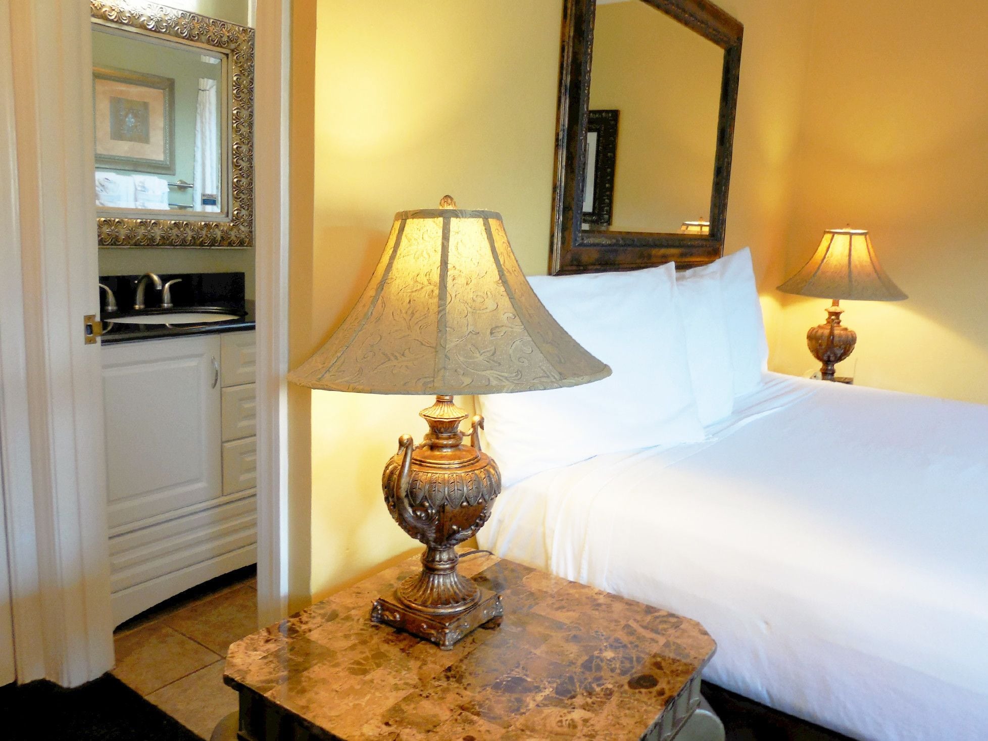 A hotel room with a bed, a nightstand with a lamp, a bathroom with a sink and mirror, and a marble-tiled floor.
