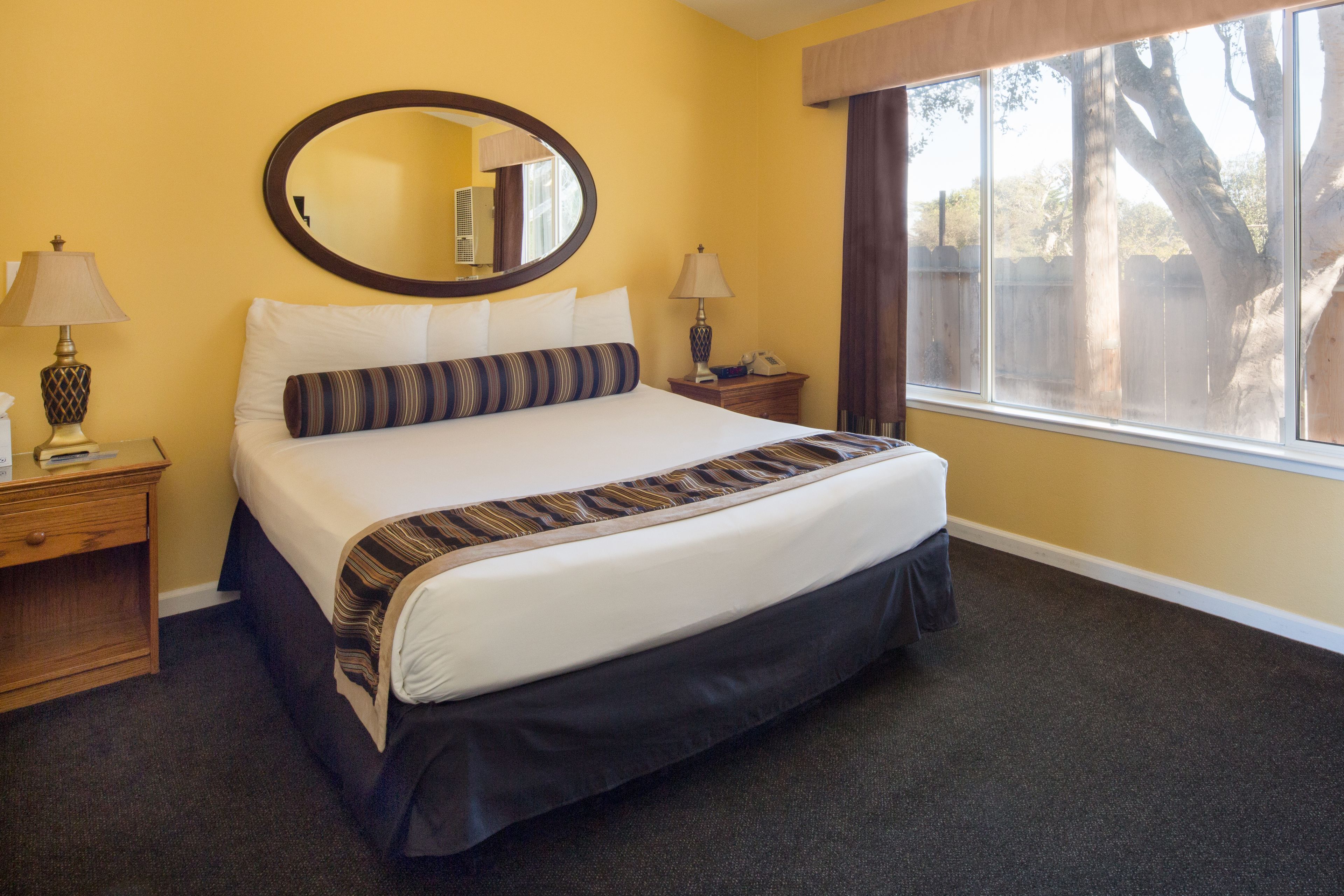 A hotel room with a large bed, two bedside tables, a round mirror, and a window with a view of trees.