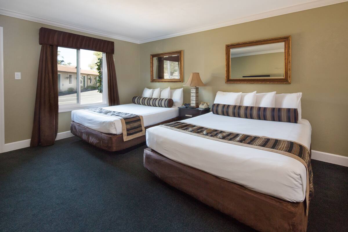 A hotel room with two beds, a nightstand, a lamp, and a large window with curtains.