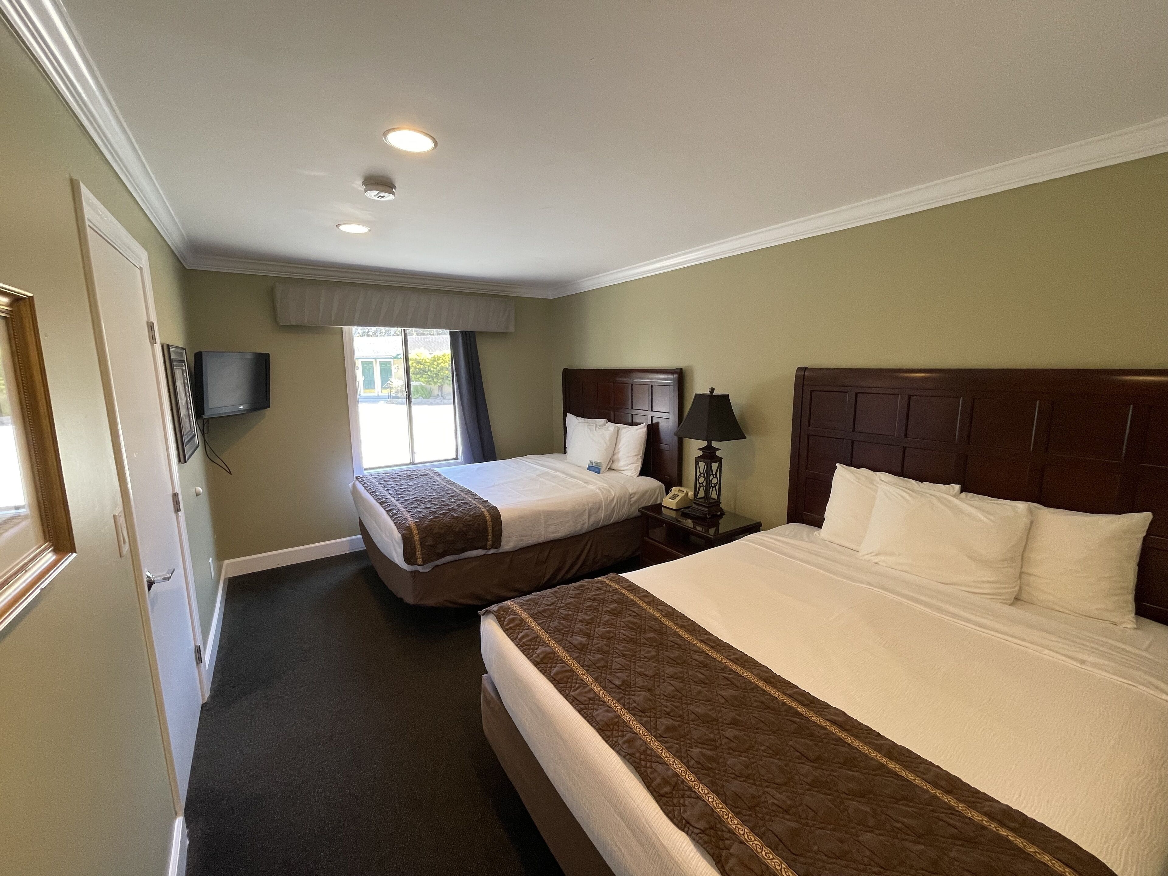 A hotel room with two beds, a television, a nightstand with a lamp, and a window with curtains.