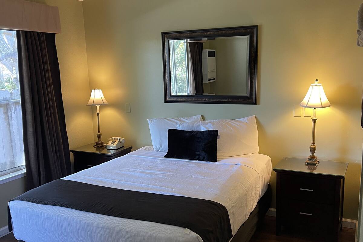 A neatly made bed with a black and white bedspread, two bedside lamps, a nightstand with a telephone, and a large mirror above the bed.