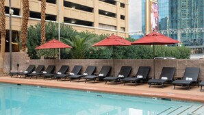 A poolside area with lounge chairs and red umbrellas, a spa pump sign, and a building with a large billboard in the background. - The Carriage House (Las Vegas)