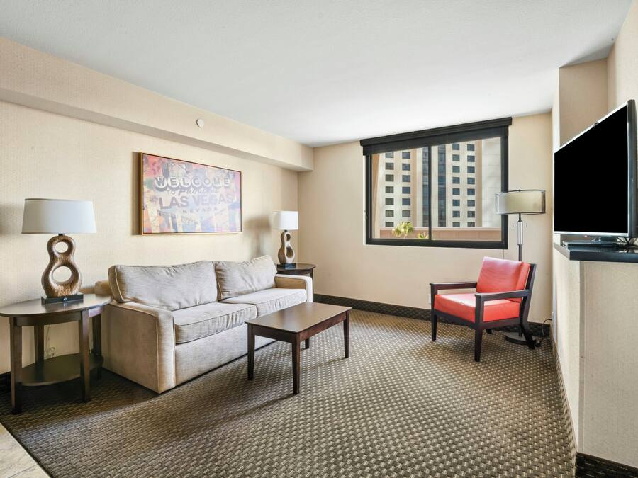 A hotel room with a sofa, a coffee table, a red chair, a TV, and a window with a city view.