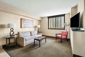 A hotel room with a sofa, a coffee table, a red chair, a TV, and a window with a city view.