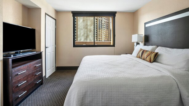 A hotel room with a bed, a TV, a dresser, and a window with blinds. - The Carriage House (Las Vegas)