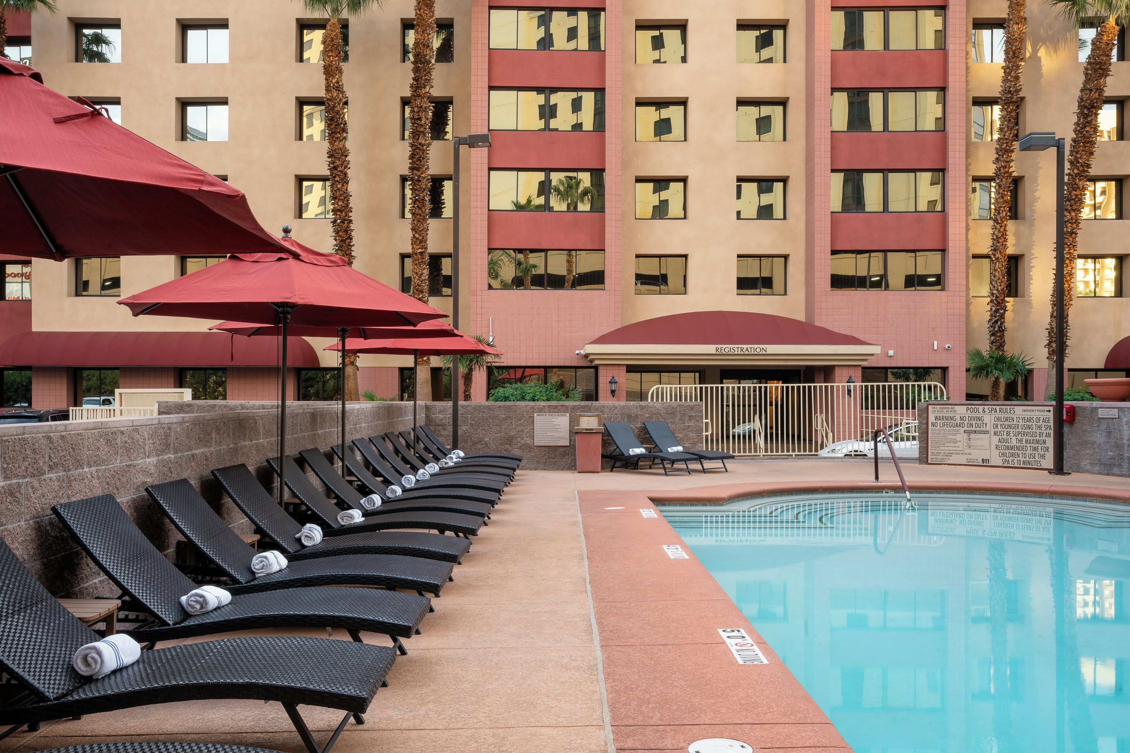 A pool area with lounge chairs and umbrellas, a registration building, and a multi-story hotel facade.