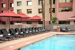 A poolside area with lounge chairs and red umbrellas, a tiled pool, and a building with multiple windows and a sign. - The Carriage House (Las Vegas)