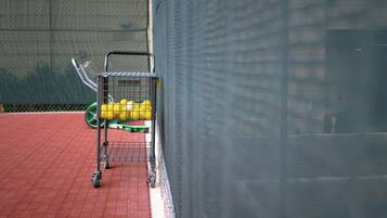 Una cancha de tenis con un carrito metálico que contiene pelotas de tenis amarillas, una bolsa de tenis verde y una raqueta de tenis.